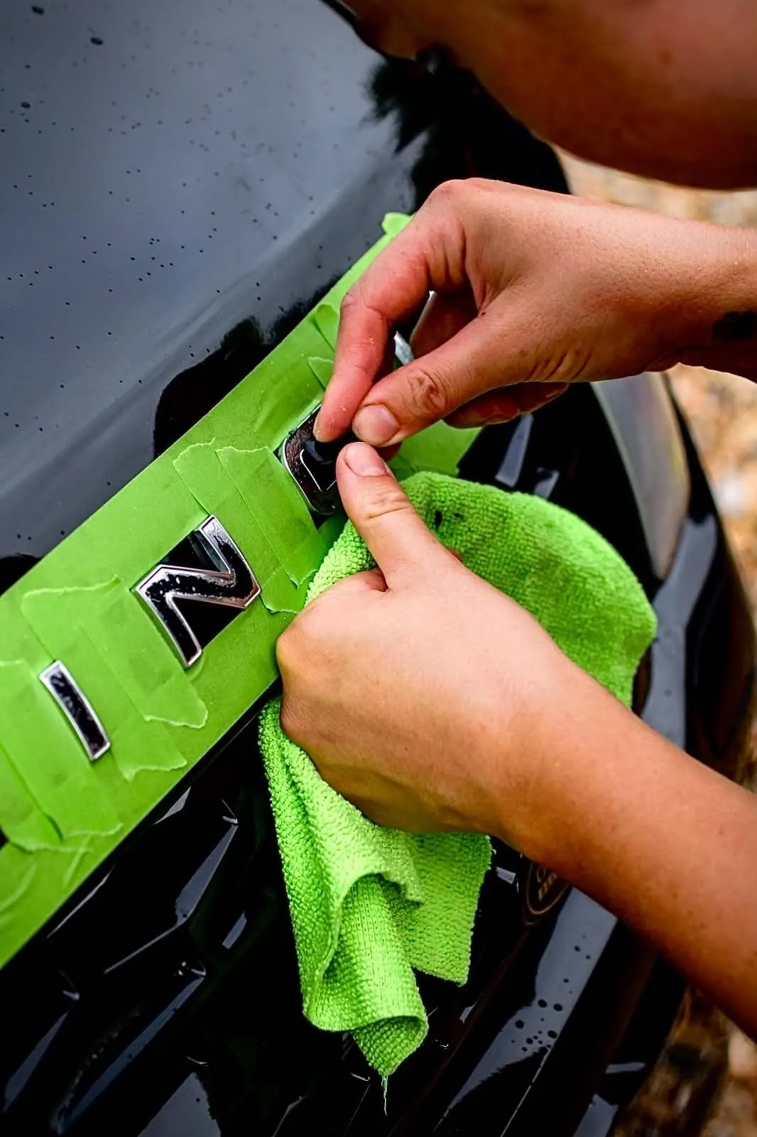 Chrome badge letters being lifted from a black bonnet, their position marked out in green tape